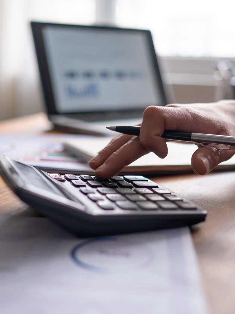 Hände tippen auf einem Taschenrechner. Im Hintergrund ein aufgeklapptes Laptop.