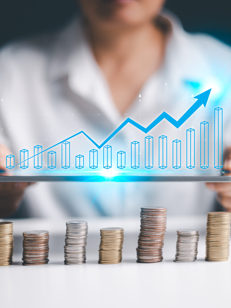 In der unteren Hälfte des Bildes: Münzstabel in verschiedenen Höhen. Darüber Hände die ein Pad halten. Darüber eien Grafik mit Säulendiagrammen und eine Kurve die Höhen sowie Tiefen und dabei eine steigende Tendenz aufweist